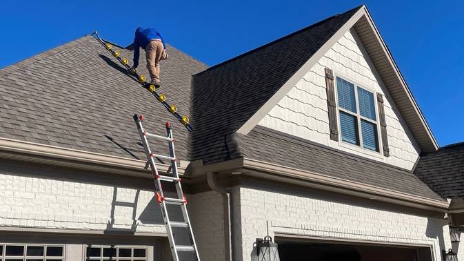 Worker on a ladder inspecting the shingle roof of a two-story house